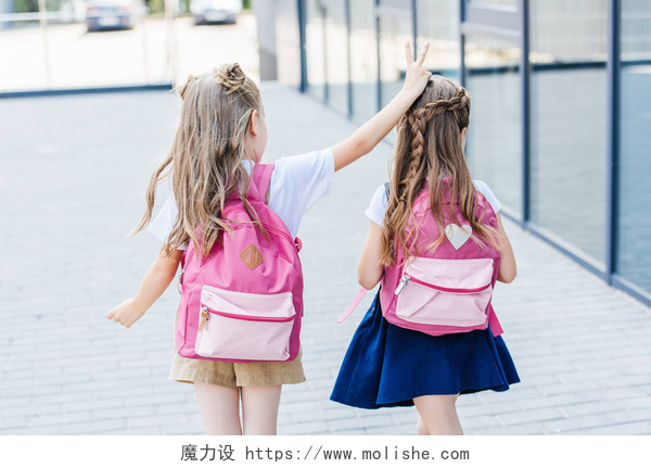 两个可爱的小女孩背着粉色的包在上学后视图的小女生做角笑话手势她的同学在街上